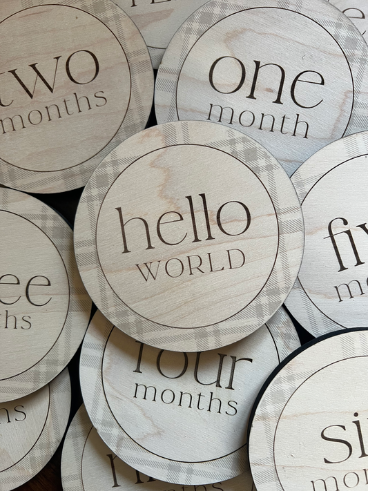 Close-up of engraved wooden baby milestone discs with plaid border and Hello World marker